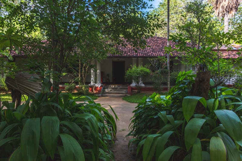 The white colonial walawwa with its red clay-tile roof glimpsed down a sand path through deep-green tropical foliage in dappled shade