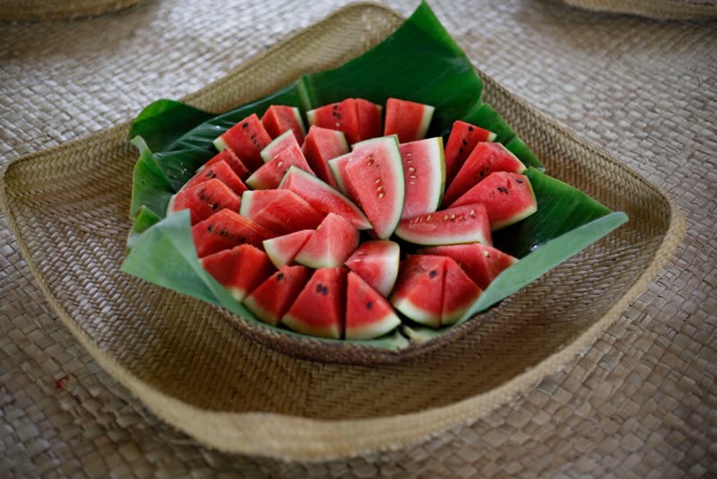 Slices of watermelon arranged on banana leaves in a woven basket resting on a straw mat.