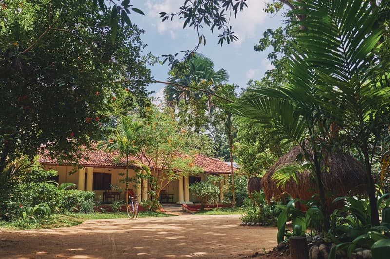 A yellow-washed pavilion building with a red roof under palms, a bicycle leaning by the pillars and a sandy track in front