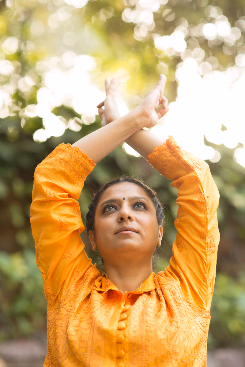 Close portrait of a woman in a saffron-orange embroidered kurta, wrists crossed overhead in a mudra, gazing up through sun-flared foliage