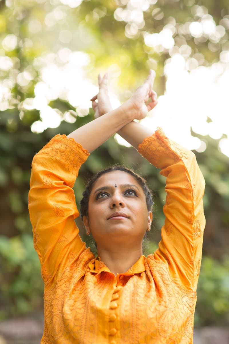 Close portrait of a woman in a saffron-orange embroidered kurta, wrists crossed overhead in a mudra, gazing up through sun-flared foliage