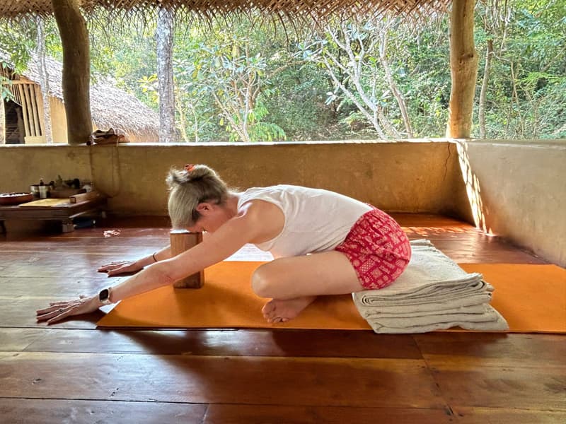 Open-sided yoga pavilion with a thatch fringe and polished wood floor: a woman stretches over a cork block on an orange mat, jungle all around